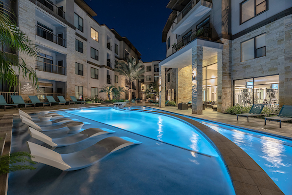Apartment courtyard pool at night with illuminated water, in-water lounge chairs, palm trees, and surrounding lounge seating near fitness-centre windows.