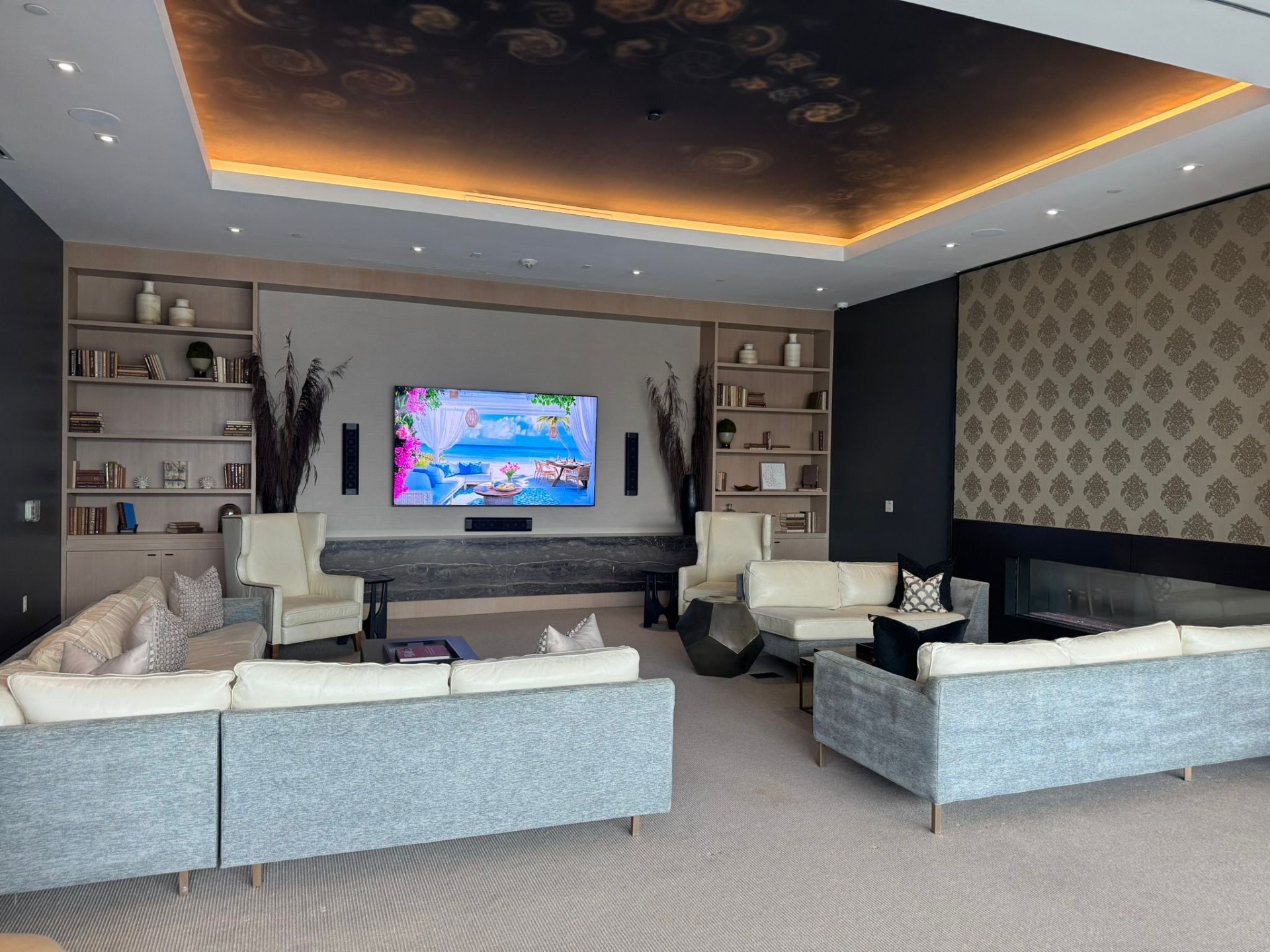 Modern lounge room with multiple sofas, wall-mounted TV, built-in shelving, decorative vases and books, and a contemporary fireplace under warm lighting.