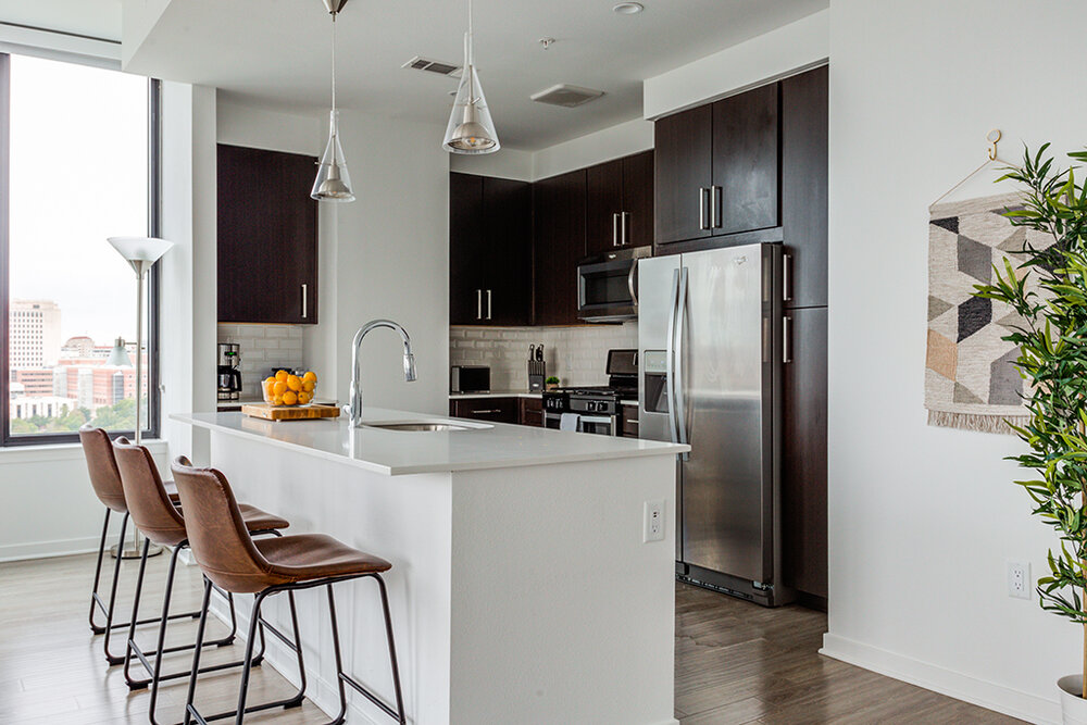 Modern apartment kitchen with dark wood cabinets, stainless steel appliances, white countertops, pendant lights, and a breakfast bar with three leather barstools.