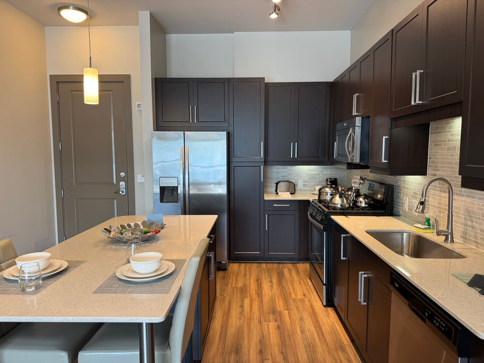 Modern apartment kitchen with dark wood cabinets, cream countertops, stainless steel appliances, a pendant light, and a table set with crockery.