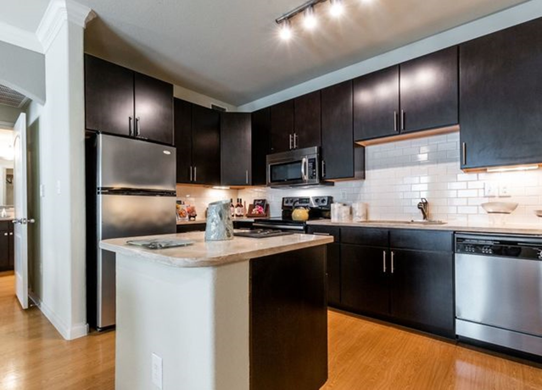Modern apartment kitchen with dark wood cabinets, stainless steel appliances, marbled countertops, and a white tiled backdrop.