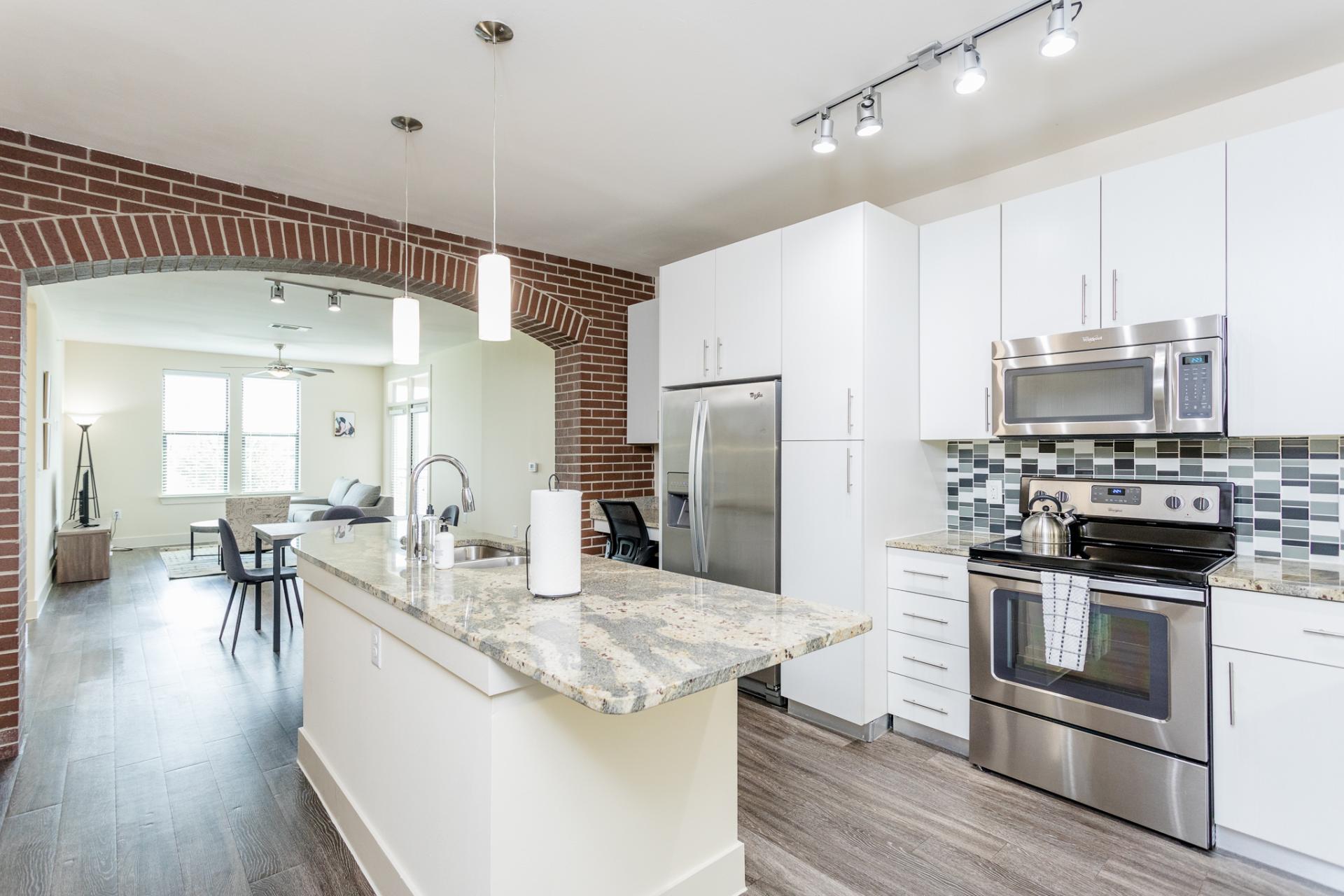 A modern open plan apartment kitchen and dining area.  With shiny, white cabinets, stainless steel appliances, marble countertops, black and white tiled backdrop, and pendant lights over the island table. A brick archway leading into living, with bright natural lighting, light coloured sofas and a TV..