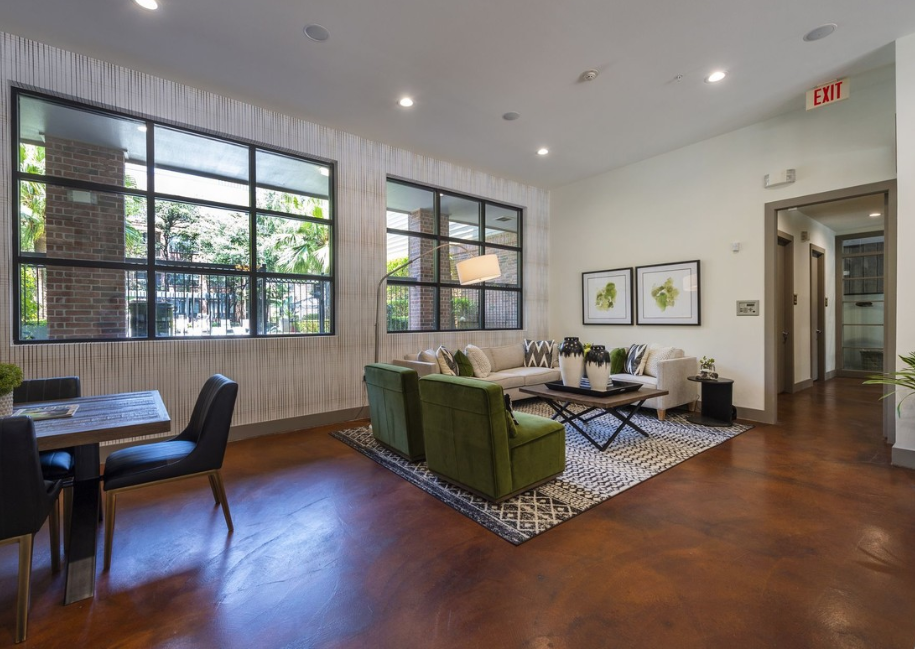 A modern lounging area decorate with plants, vases and art work. Lots of windows letting natural light in. A wooden-looking shiny floor, with a pattern rug, cream coloured sofas, and green chairs. A table area with black leather chairs. A view of a corridor and door ways.