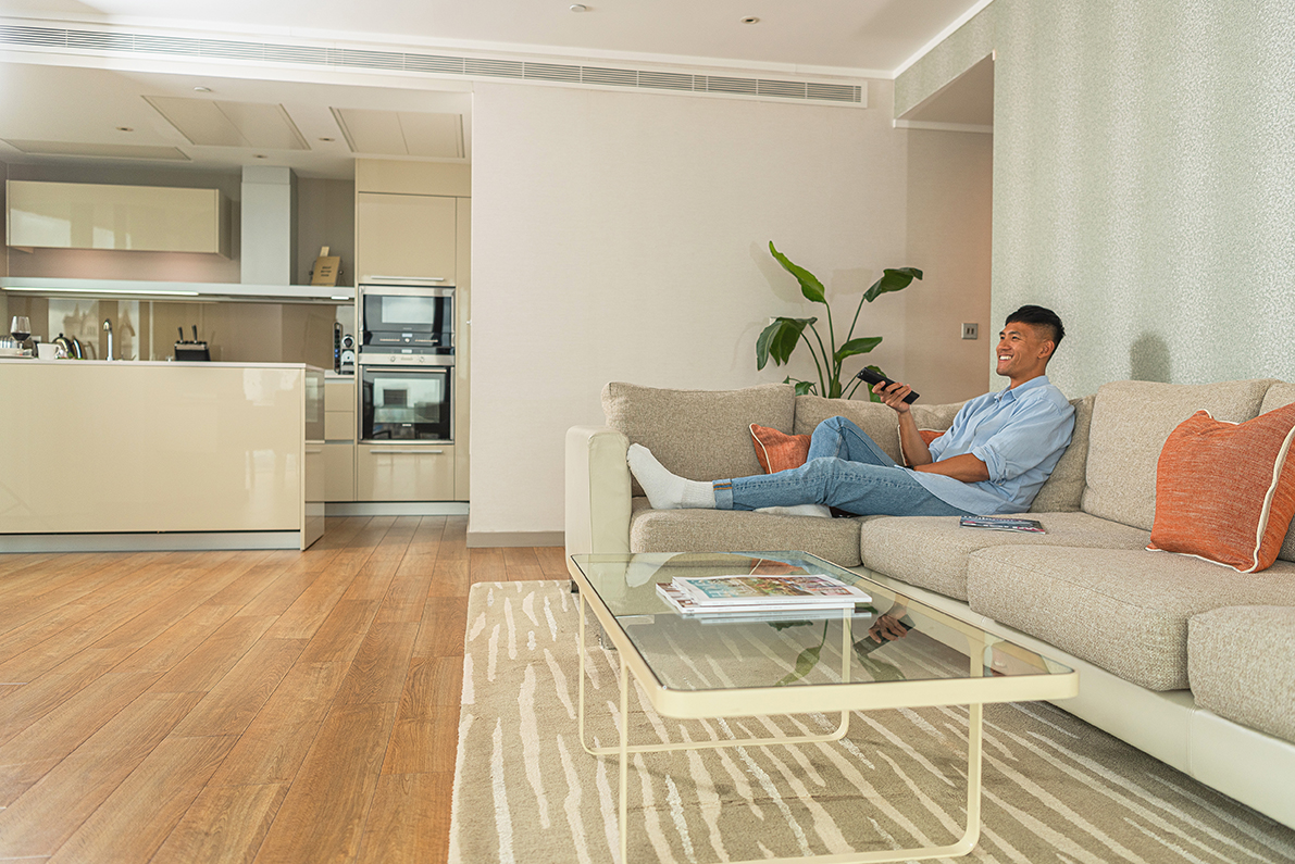 An image of a man in a Serviced Apartment displaying a captivating blend of modernity and comfort, where the sleek, white-walled sanctuary is centered around a statement-making sofa. The glass coffee table and stainless steel kitchen accentuate the room's minimalist elegance, creating a cozy yet luxurious haven for filmmakers and creatives.