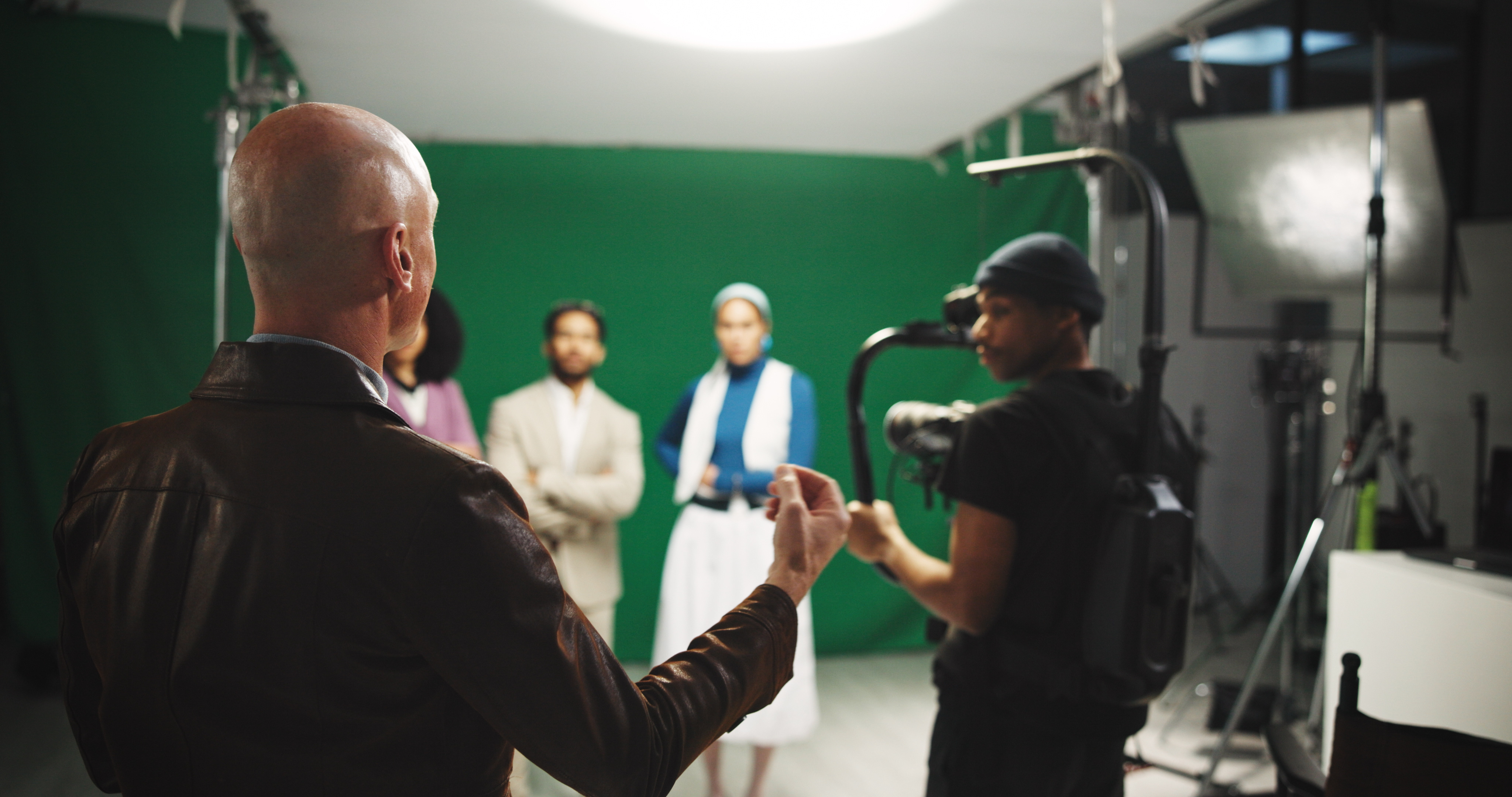 Film director guiding a cameraman on a studio set, with actors performing in front of a green screen in the background.