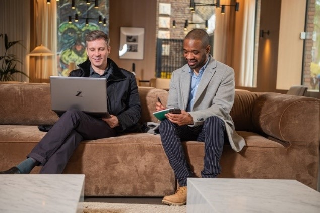 Two men sat on a sofa, one with a laptop, one writing notes.