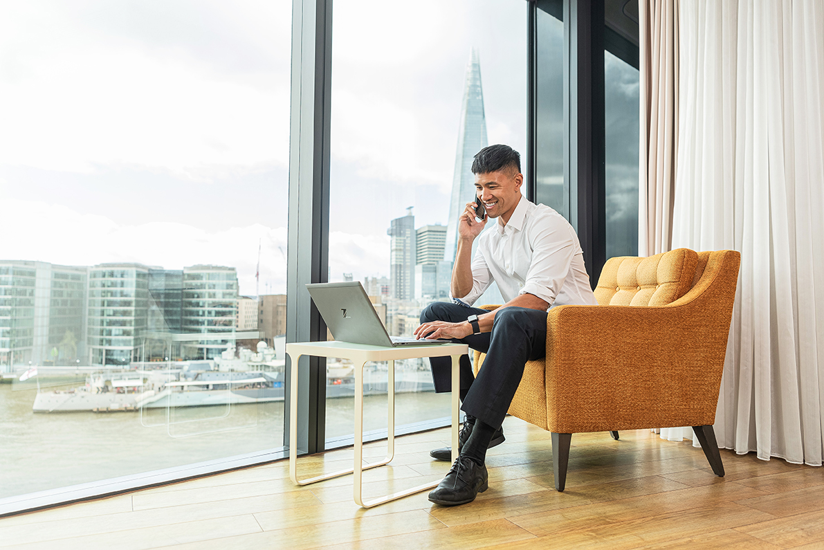 A man sat on a chair, taking a phone call, smiling. London city in the background through the window.