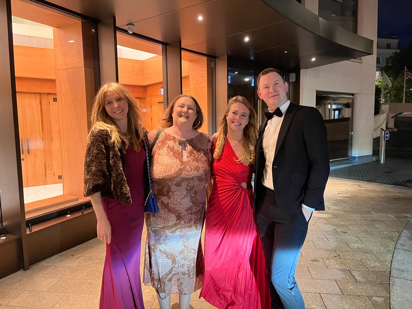 Four people dressed in formal evening wear smiling outside a building entrance at night, with warm lighting reflecting off the wooden doors behind them.