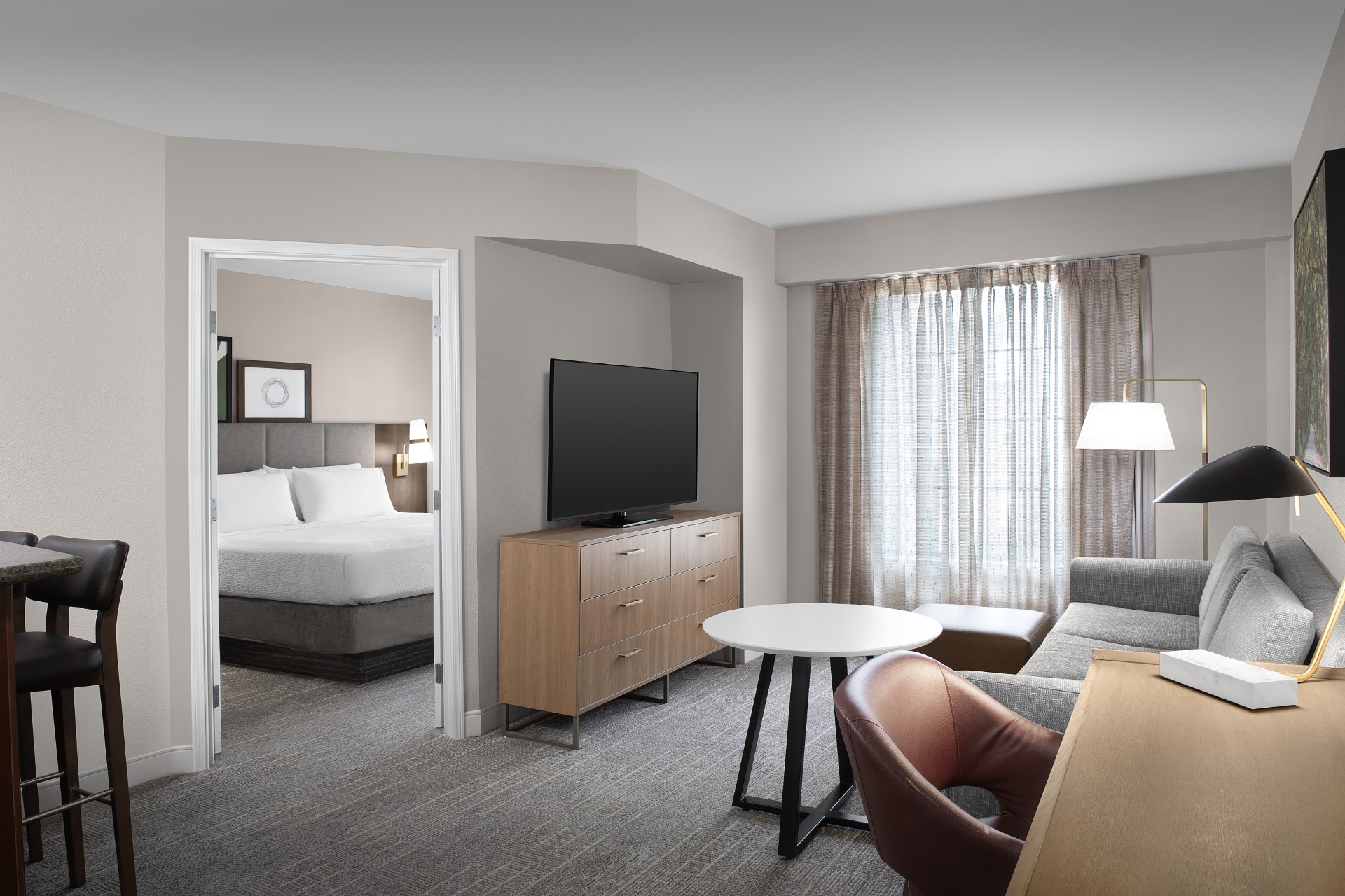 Modern hotel suite with a separate bedroom, flat-screen TV on a wooden dresser, gray sofa, desk, and large window with sheer curtains.