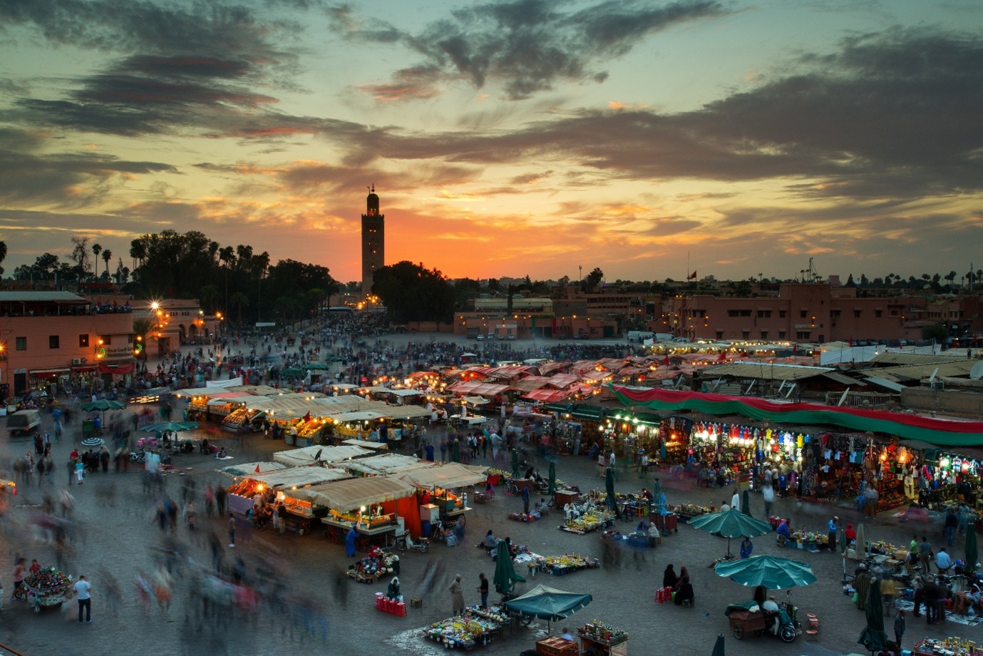 Sunset over a bustling market square with food stalls and crowds, mosque minaret in the background.