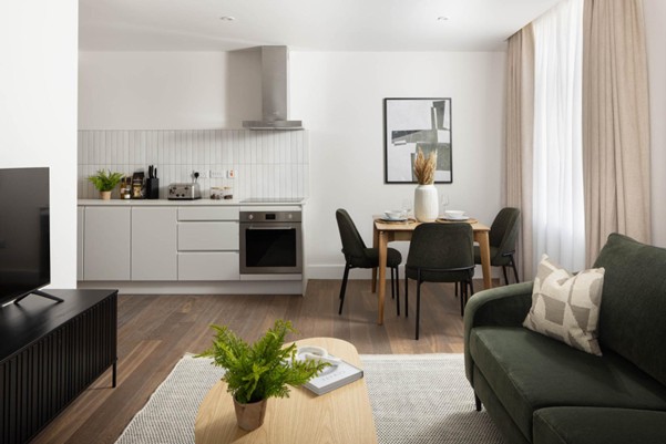 A modern open-plan living space with a small kitchen, dining table for four, green sofa, and light wood furniture.