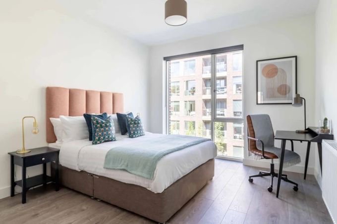 A bright bedroom with a large bed, peach upholstered headboard, desk with chair, and a window overlooking nearby buildings.