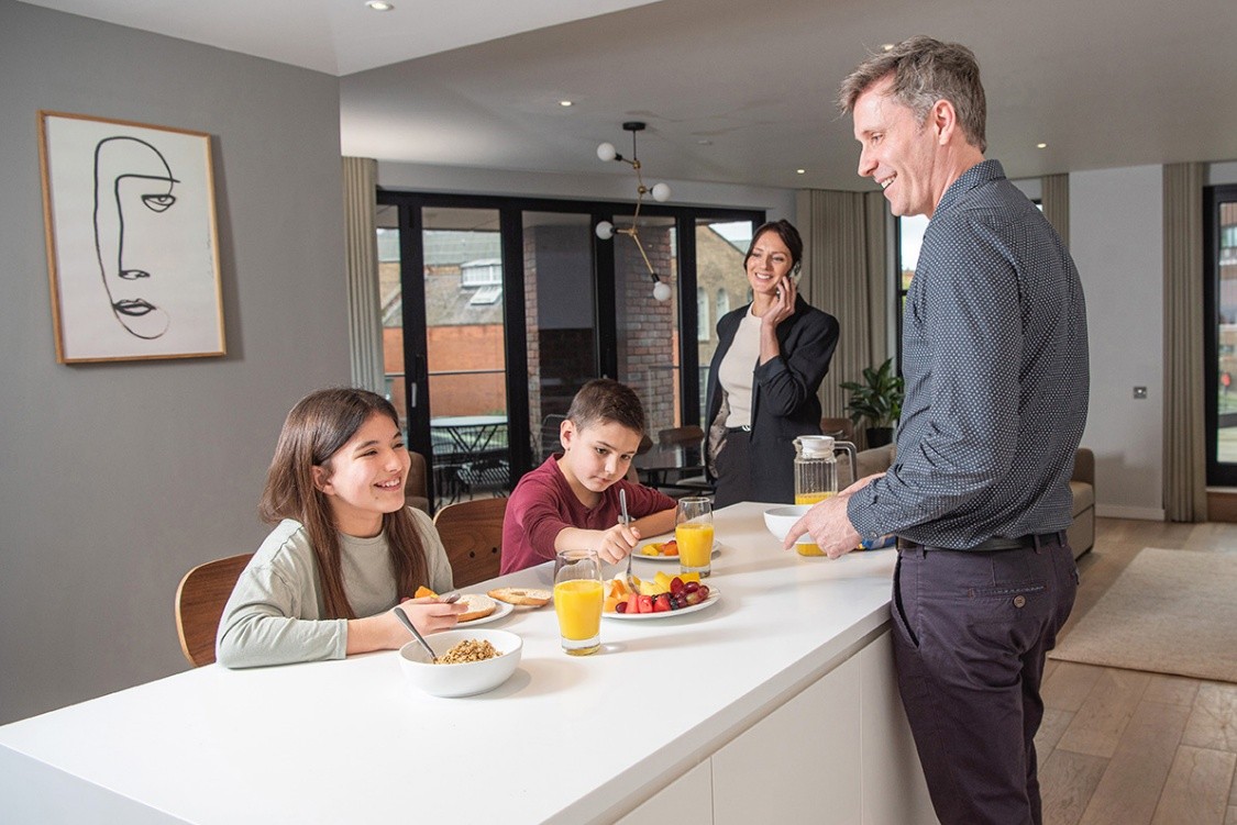 A family of four, the children sat on an island table eating breakfast. The dad smiling and mum on the phone and smiling